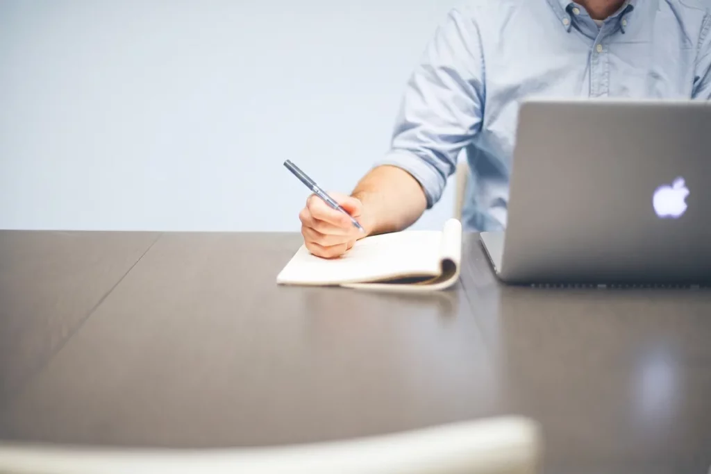Uomo che prende appunti su un quaderno accanto al portatile, il momento della scelta fra le edizioni Zoho