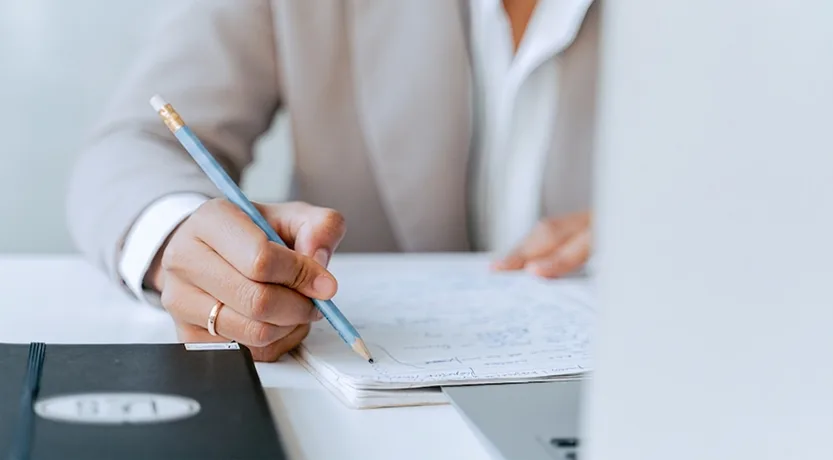 Mano che prende appunti a matita su un quaderno accanto al portatile, le voci di spesa che restano fuori dal preventivo