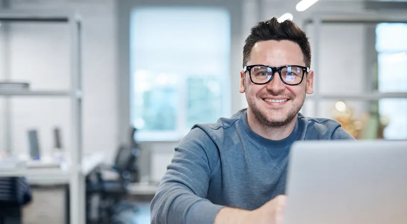 Uomo con gli occhiali sorride davanti al portatile in ufficio, copertina dell'articolo sui costi di Zoho CRM