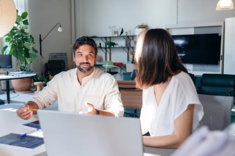 Due colleghi seduti al tavolo di un ufficio luminoso si confrontano davanti a un laptop e a documenti stampati